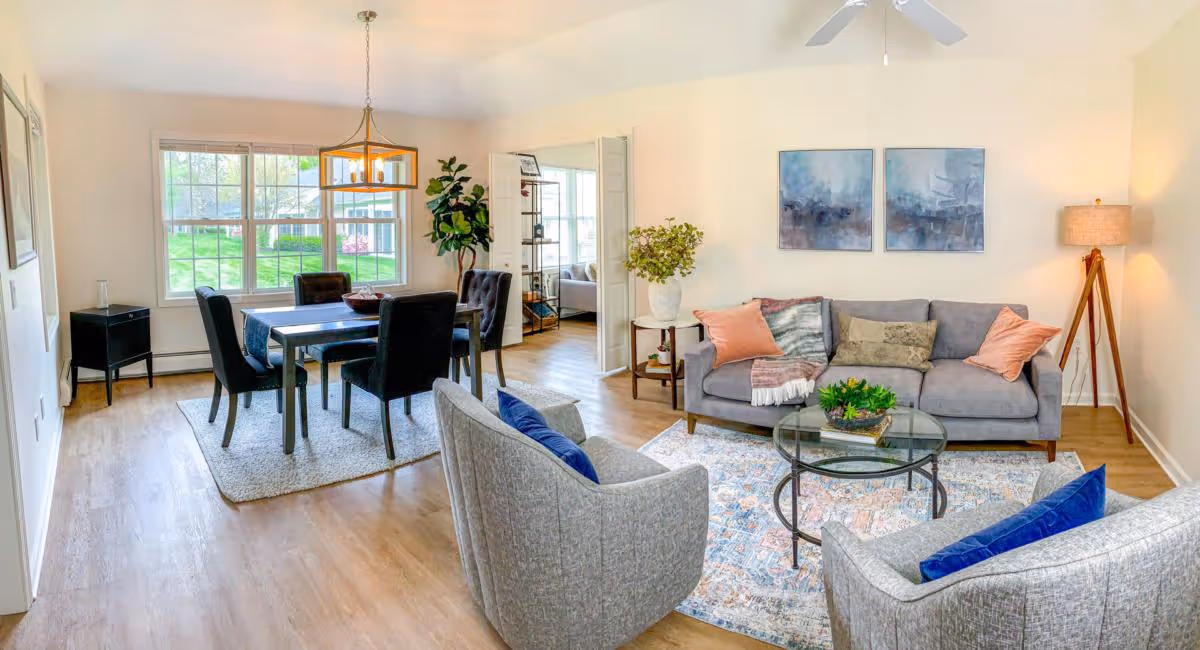 Bright open living and dining area with a sofa, two armchairs, glass coffee table, and a dining table by a large window.