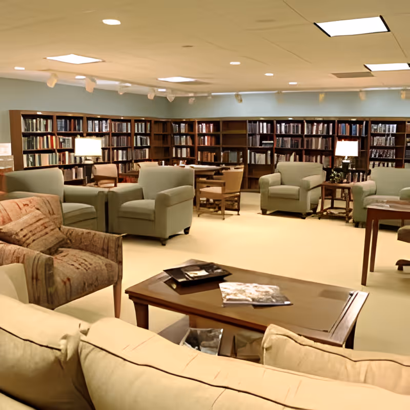 A spacious and well-lit library or reading room with multiple bookshelves filled with books along the walls. The room features several comfortable armchairs and sofas arranged around wooden coffee tables, with table lamps providing additional lighting. The carpeted floor and ceiling lights create a cozy and inviting atmosphere.