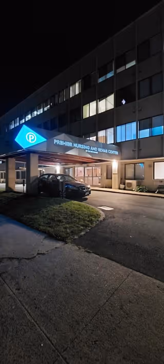 Night view of the front entrance of Premier Nursing and Rehab Center of Far Rockaway, showing a multi-story building with illuminated windows and a covered drop-off area with a car parked underneath.