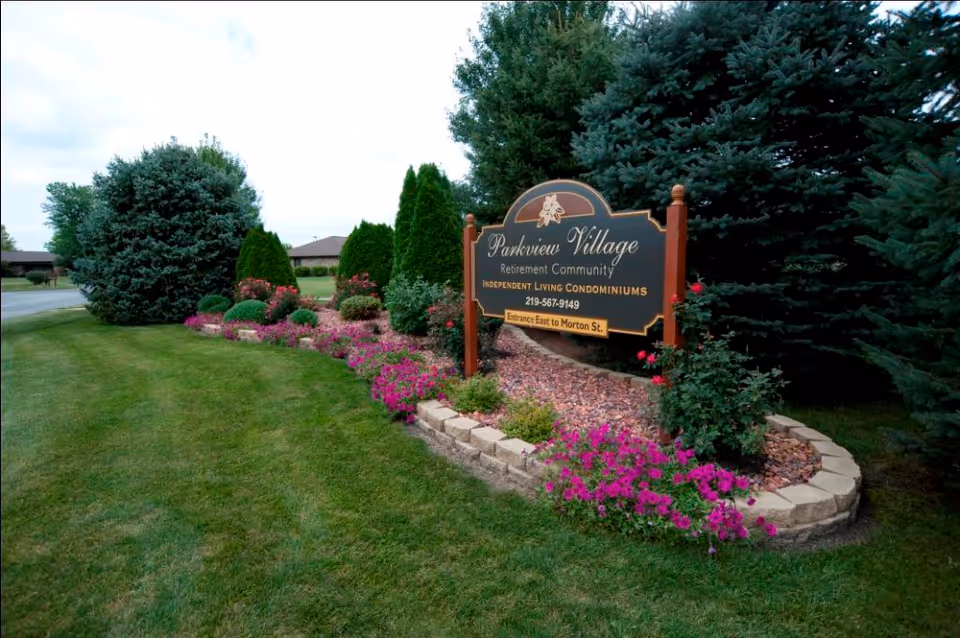 A landscaped outdoor area with a green lawn, various bushes, and pink flowers surrounding a sign that reads 'Parkview Village Retirement Community Independent Living Condominiums 219-567-9149 Entrance East to Morton St.'