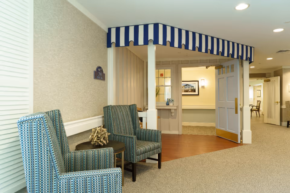 A cozy indoor seating area with two patterned armchairs and a small round table between them, placed near a hallway entrance with a blue and white striped awning above. The hallway has light-colored walls, carpeted floors, and an open door leading to another room with chairs and tables visible in the background.