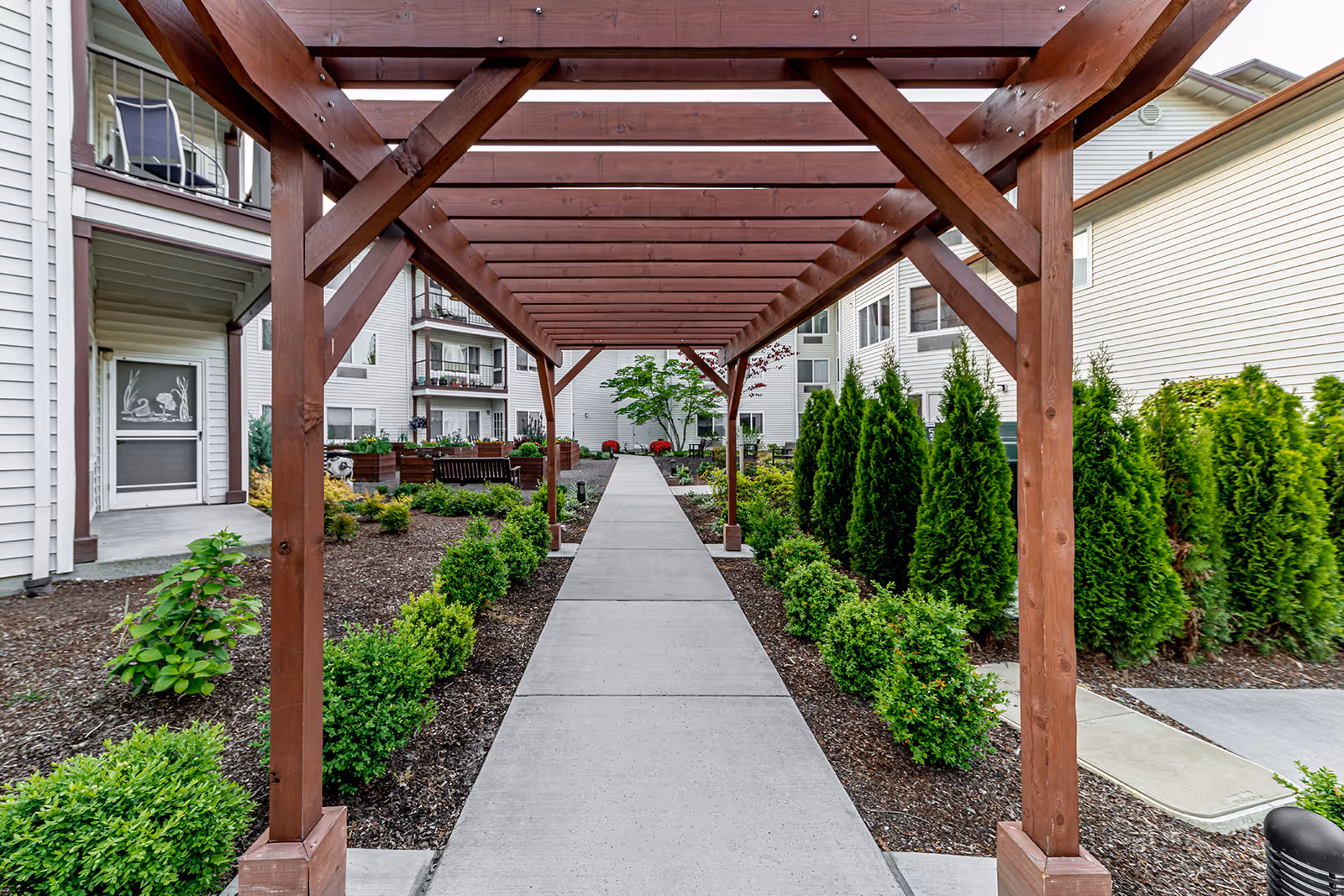 A concrete pathway runs through a garden area with neatly trimmed bushes and small trees on either side. A wooden pergola structure spans over the pathway. In the background, there are white residential buildings with balconies and windows. There are benches and raised garden beds along the path.