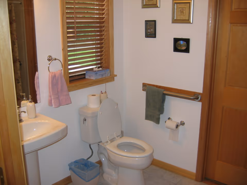 A small bathroom with a white toilet with the lid open, a pedestal sink with soap dispenser, a window with wooden blinds, a pink hand towel hanging on a ring, a green towel hanging on a grab bar, a toilet paper holder with a roll of toilet paper, a small trash bin, and framed pictures on the wall.
