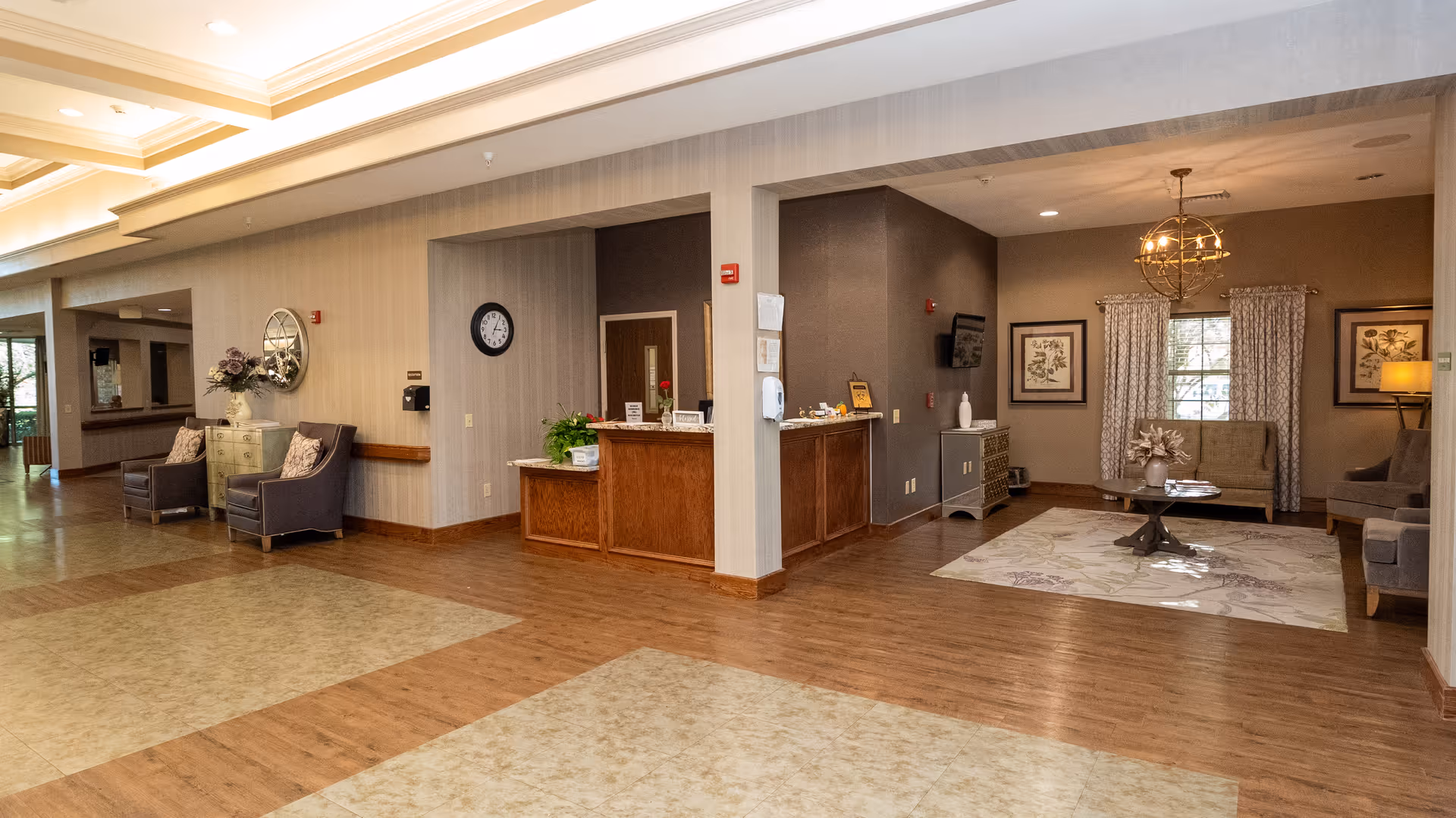 Interior view of a healthcare facility lobby area with a reception desk in the center, seating areas with chairs and a small table with flowers on the left, and a cozy sitting area with a sofa, armchairs, a coffee table, and framed artwork on the walls to the right. The floor is a combination of wood and tile, and the space is well-lit with ceiling lights and a decorative hanging light fixture.
