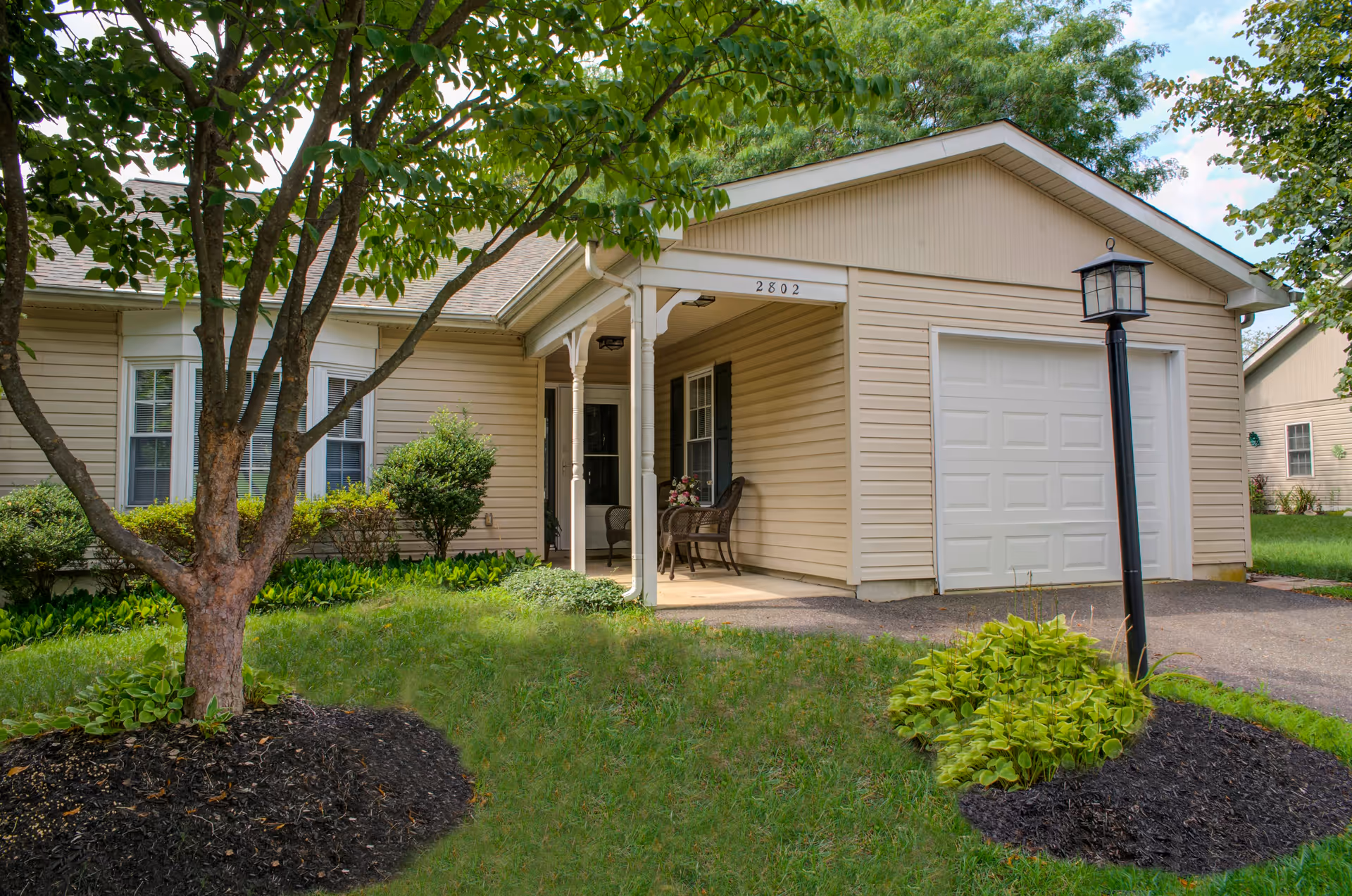 Exterior view of a single-story beige house with a small covered porch, a white garage door, and a lamp post in the front yard. There are green bushes, a tree, and a well-maintained lawn surrounding the house.