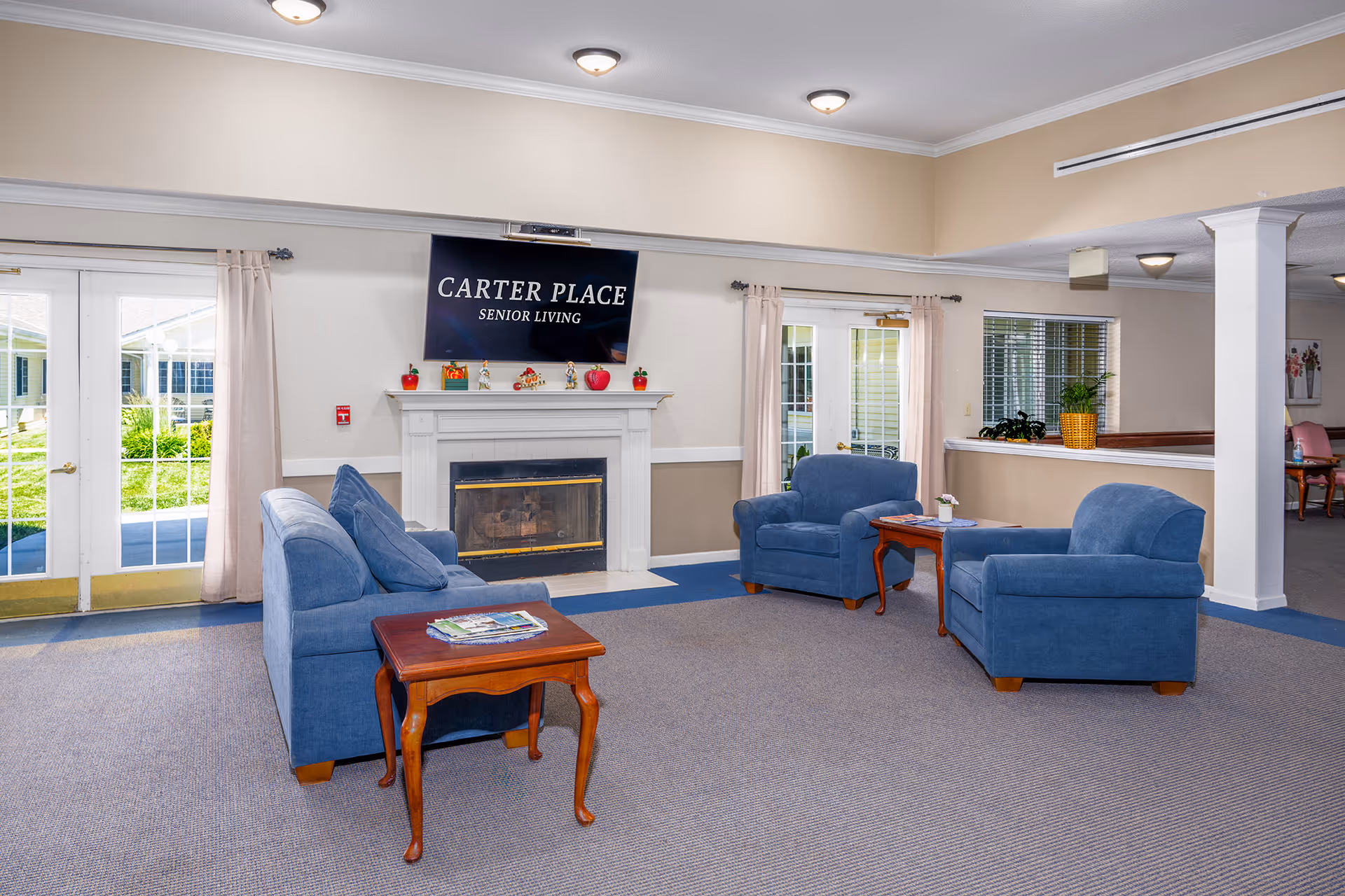 A cozy senior living common area with blue upholstered chairs and a matching sofa arranged around wooden side tables. A white fireplace with decorative items on the mantel is centered on the wall, above which is a large screen displaying 'Carter Place Senior Living'. French doors with light curtains lead outside, and there are windows with blinds and plants on the right side.
