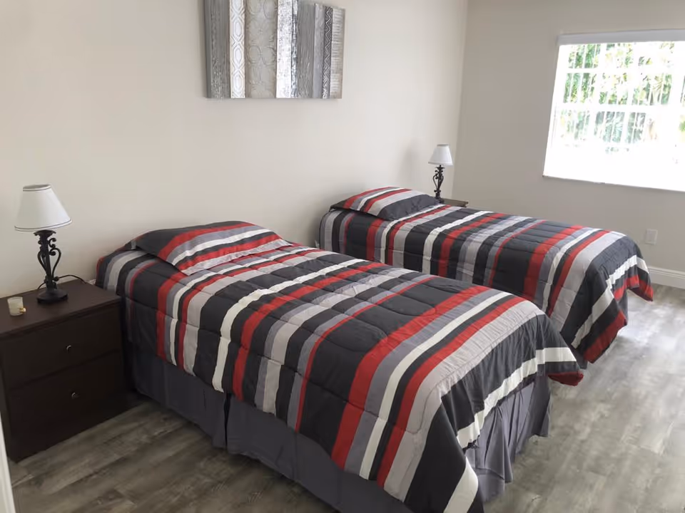 Two twin beds with red, gray, black, and white striped bedding in a bright, minimally furnished bedroom with nightstands and lamps.