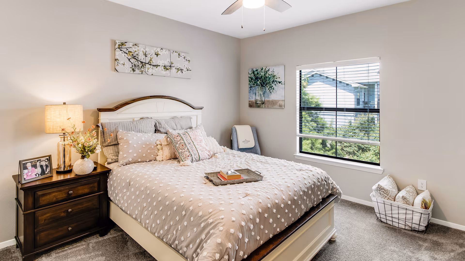 A cozy bedroom with a large bed featuring a beige and white textured comforter and multiple decorative pillows. A wooden nightstand with a lamp, a vase of flowers, and framed photos sits beside the bed. The room has light gray walls with floral artwork, a window with blinds showing greenery outside, a ceiling fan, and a basket with pillows on the carpeted floor.