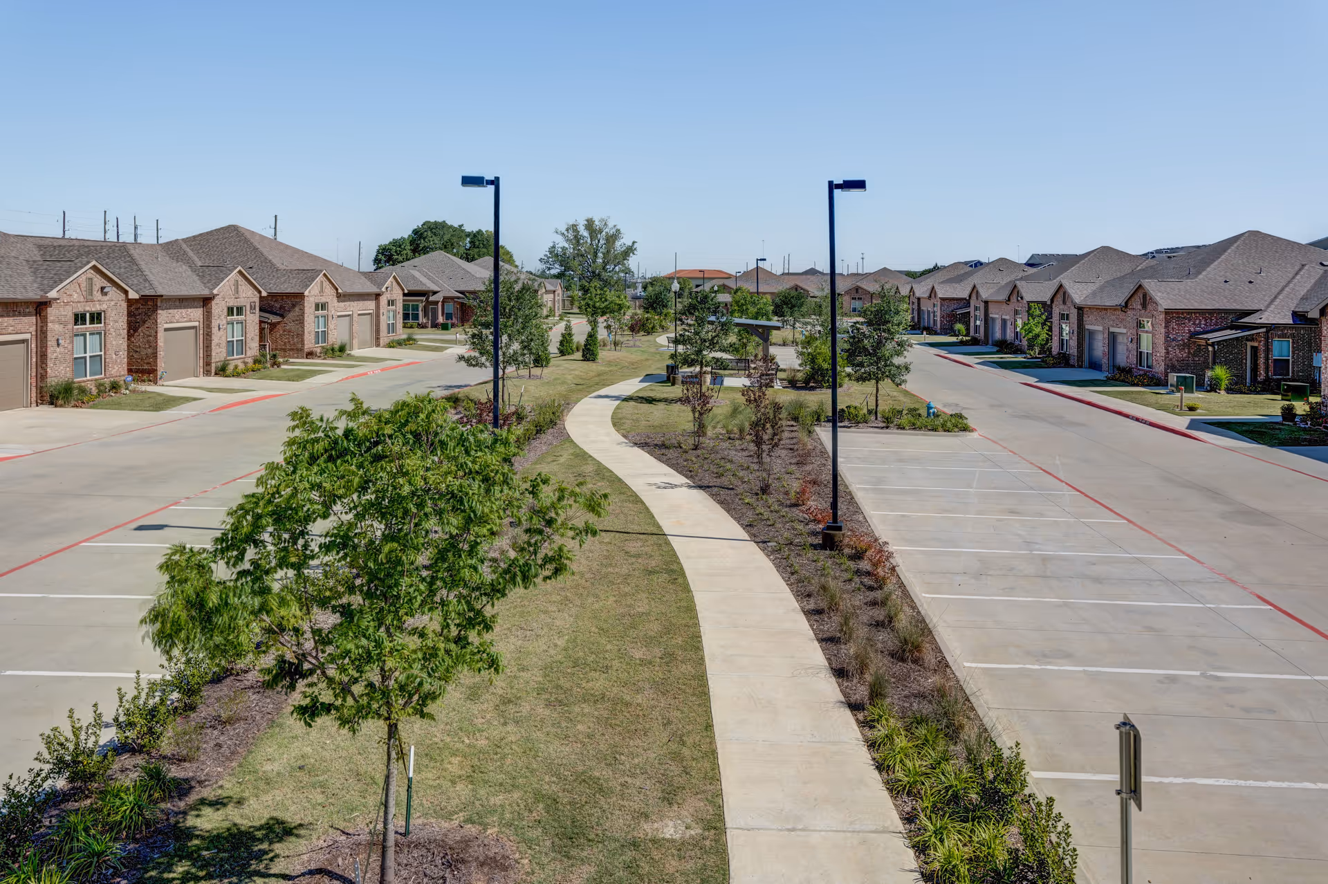 A wide paved walkway flanked by young trees and landscaping runs through the center of a residential community with single-story brick buildings on either side. The buildings have garages and driveways, and there are parking spaces along the road. The sky is clear and blue.