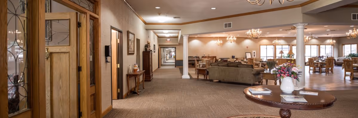 Wide hallway in a senior living facility with carpeted floor, beige walls, and wooden doors on the left. The hallway leads to a spacious common area with multiple chandeliers, sofas, armchairs, and dining tables. A round wooden table with a white vase of flowers and some brochures is in the foreground on the right.