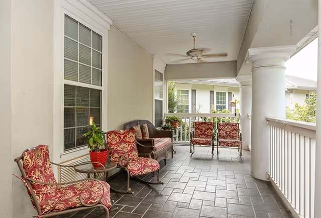 Covered outdoor porch with patterned cushioned chairs, a wicker loveseat, potted plants, a ceiling fan, and tiled floor overlooking a railing.