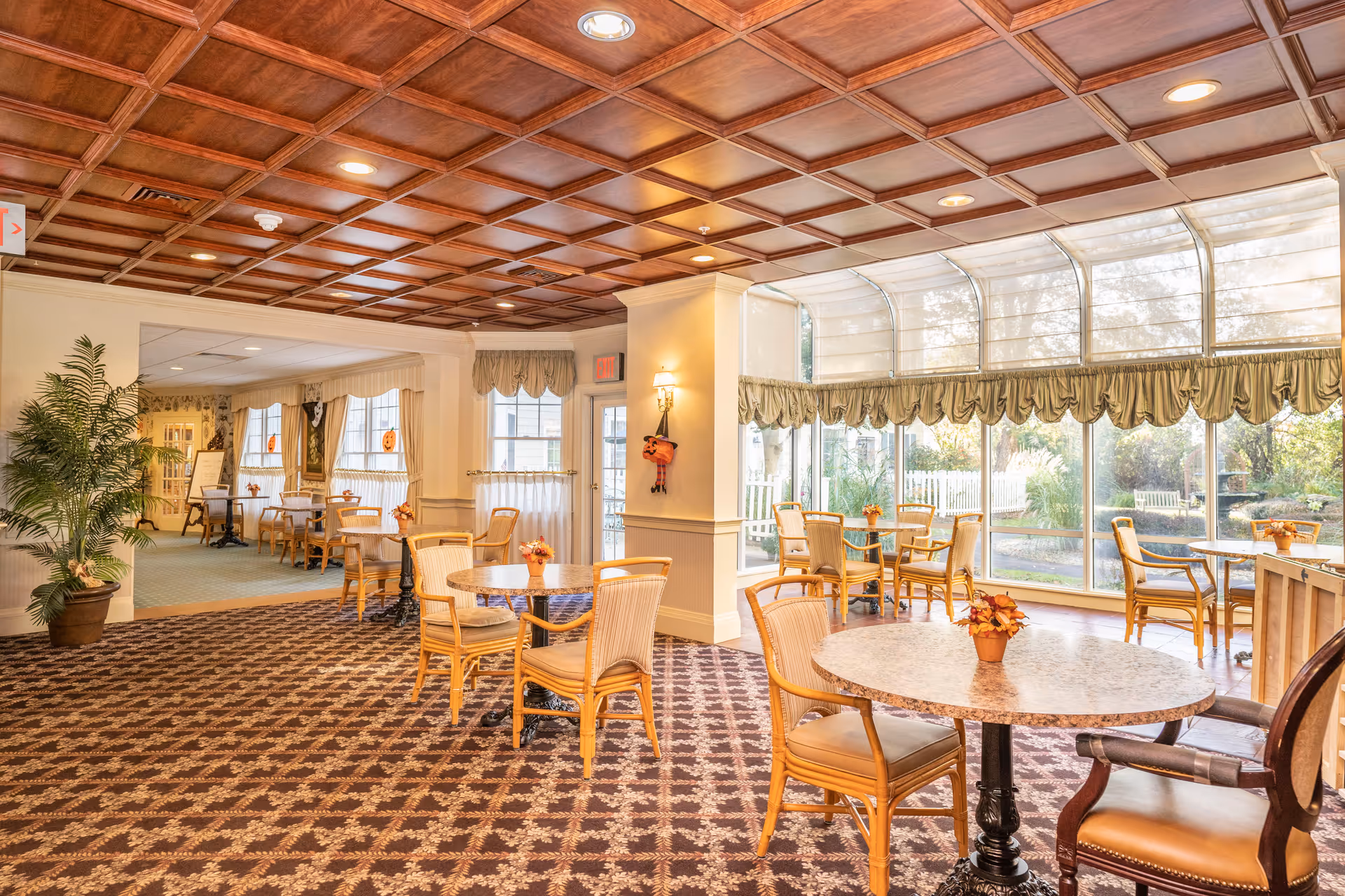 A bright and spacious dining area in an assisted living community with round tables and wooden chairs. The room features a patterned carpet, a coffered wooden ceiling with recessed lighting, large windows with valances allowing natural light to fill the space, and small flower arrangements on each table. There is a view of a garden outside through the windows.