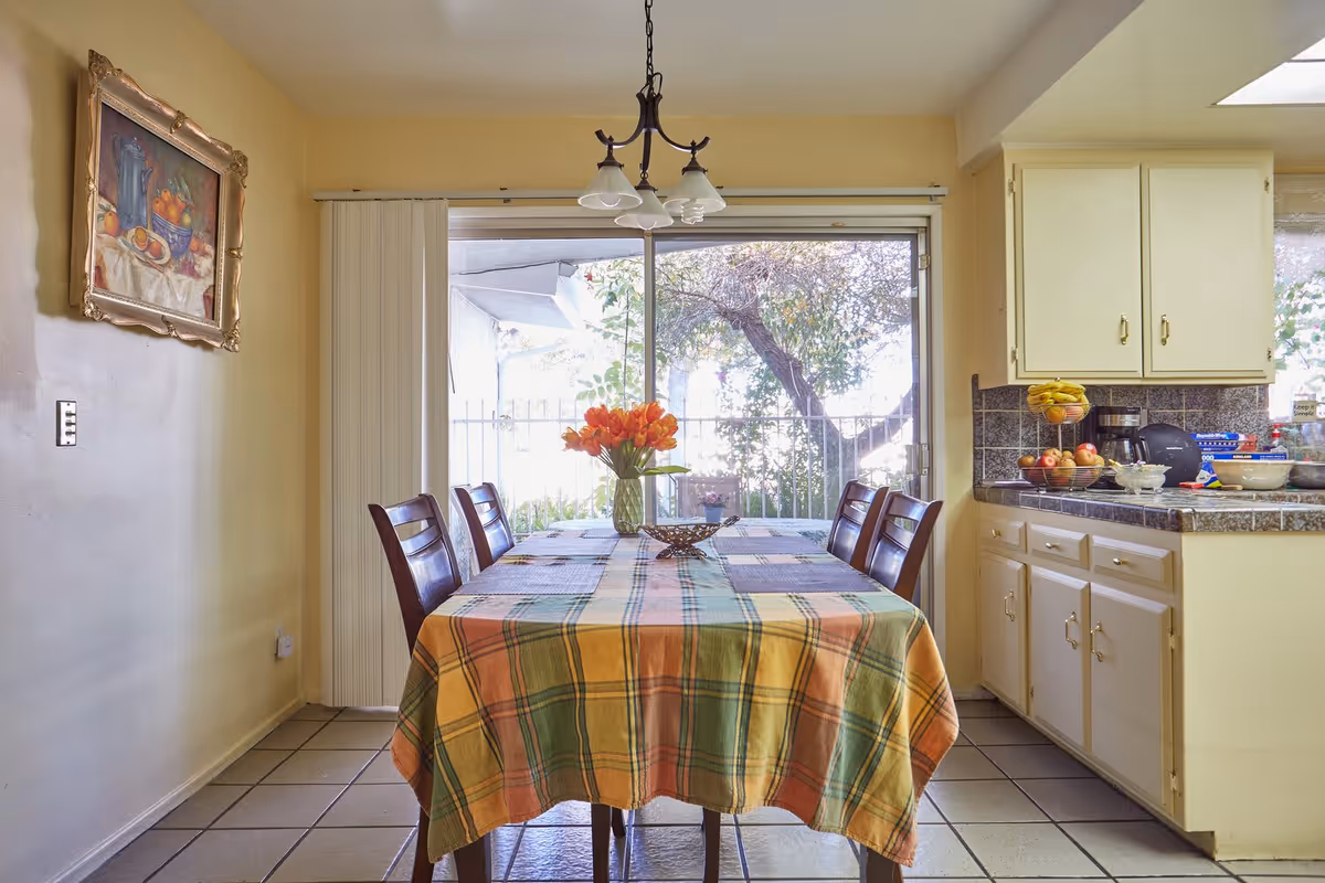 A dining area with a rectangular table covered by a colorful plaid tablecloth. Four wooden chairs surround the table. A vase with orange flowers is placed in the center of the table. Behind the table is a large sliding glass door with vertical blinds, leading to an outdoor area with trees. To the right, there is a kitchen counter with cabinets above and below, holding various fruits and kitchen items. A framed painting hangs on the left wall.