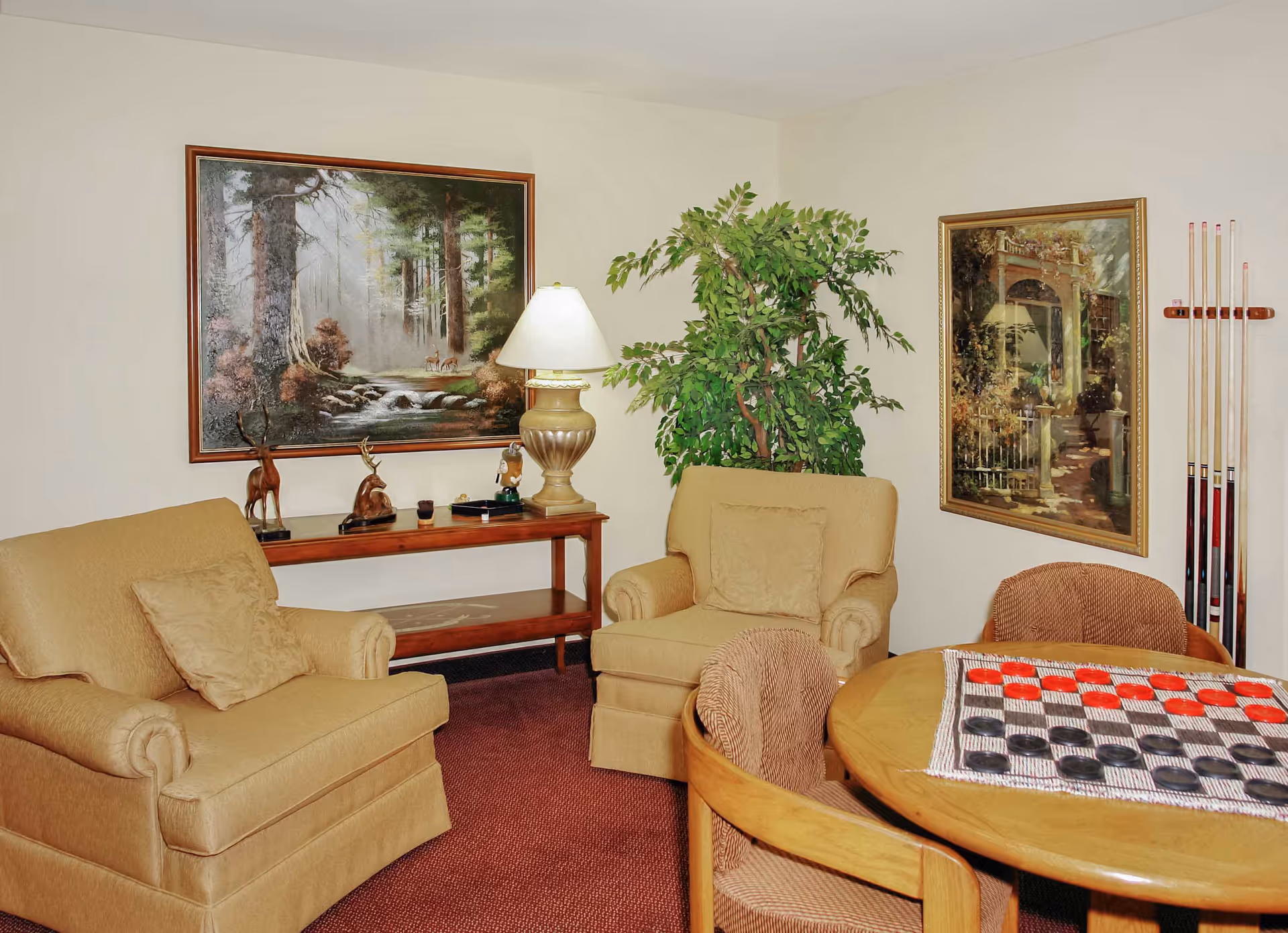 A cozy living room area with two beige armchairs, a wooden table with a checkers game set up, and a wooden console table with decorative items including two deer figurines and a lamp. There are two framed paintings on the walls and a large green potted plant in the corner.