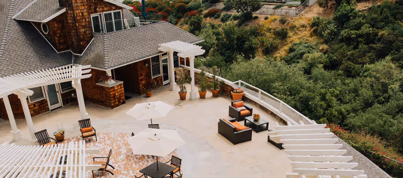 Outdoor patio area at Crown Cove featuring seating arrangements with chairs, tables, and umbrellas. The patio is surrounded by white pergolas and overlooks a lush green hillside.