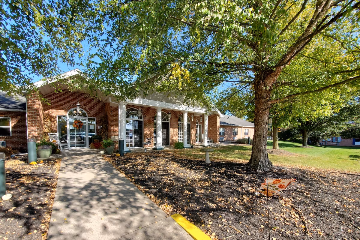 Front exterior view of a brick building with white columns and large arched windows and doors. A concrete walkway leads to the entrance, surrounded by landscaped areas with mulch, plants, and a large tree with green leaves. The sky is clear and blue.