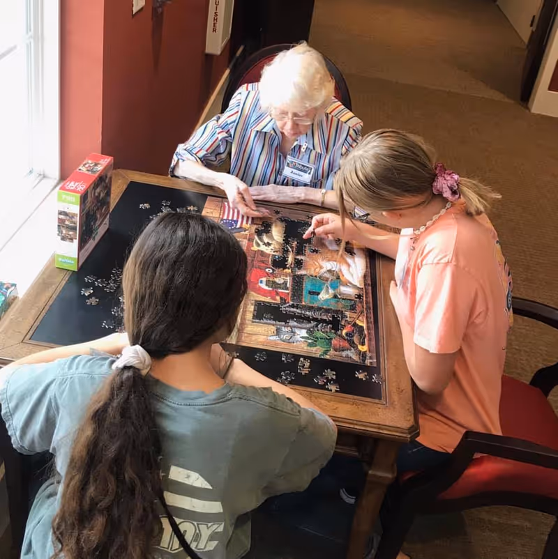 An elderly woman and two young girls sitting around a wooden table working together on assembling a jigsaw puzzle near a window in a well-lit room.