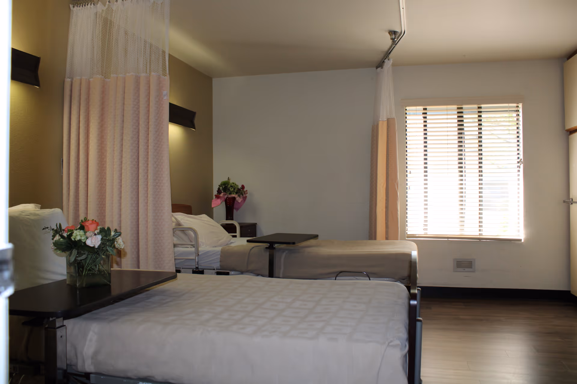 A room with two hospital-style beds separated by privacy curtains. Each bed has a small overbed table, and there are flower arrangements on the tables and a nightstand. A window with blinds allows natural light into the room, which has wood flooring and neutral-colored walls.