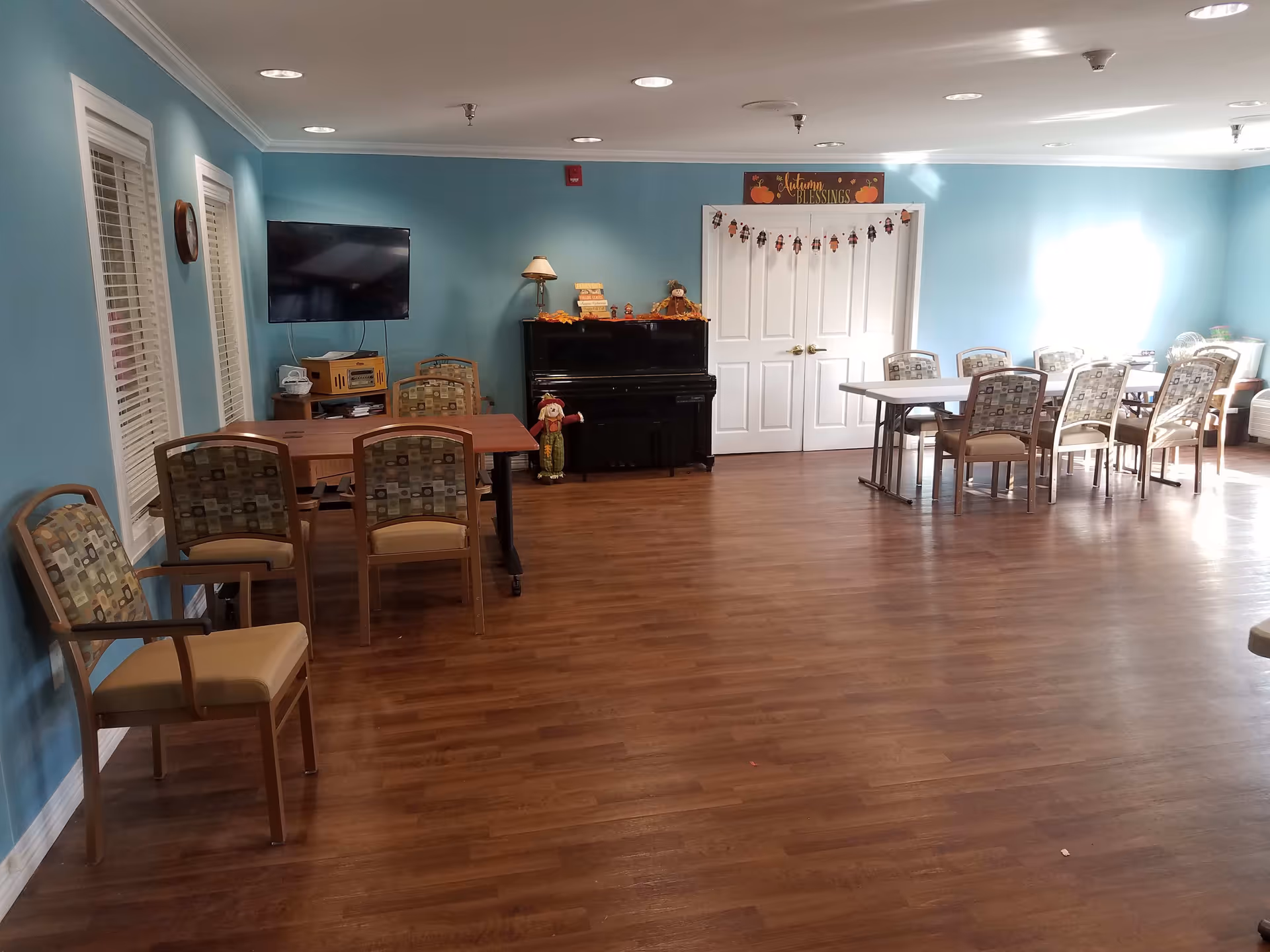 A spacious room with light blue walls and wood flooring featuring two sets of tables and chairs. On the left side, there is a wall-mounted flat-screen TV above a small cabinet with a stereo system. A black upright piano is positioned against the far wall, decorated with autumn-themed items and a small scarecrow figure. Above the double white doors at the back, a sign reads 'Autumn Blessings' with pumpkin decorations. The room is well-lit with ceiling lights and natural light coming from the right side.
