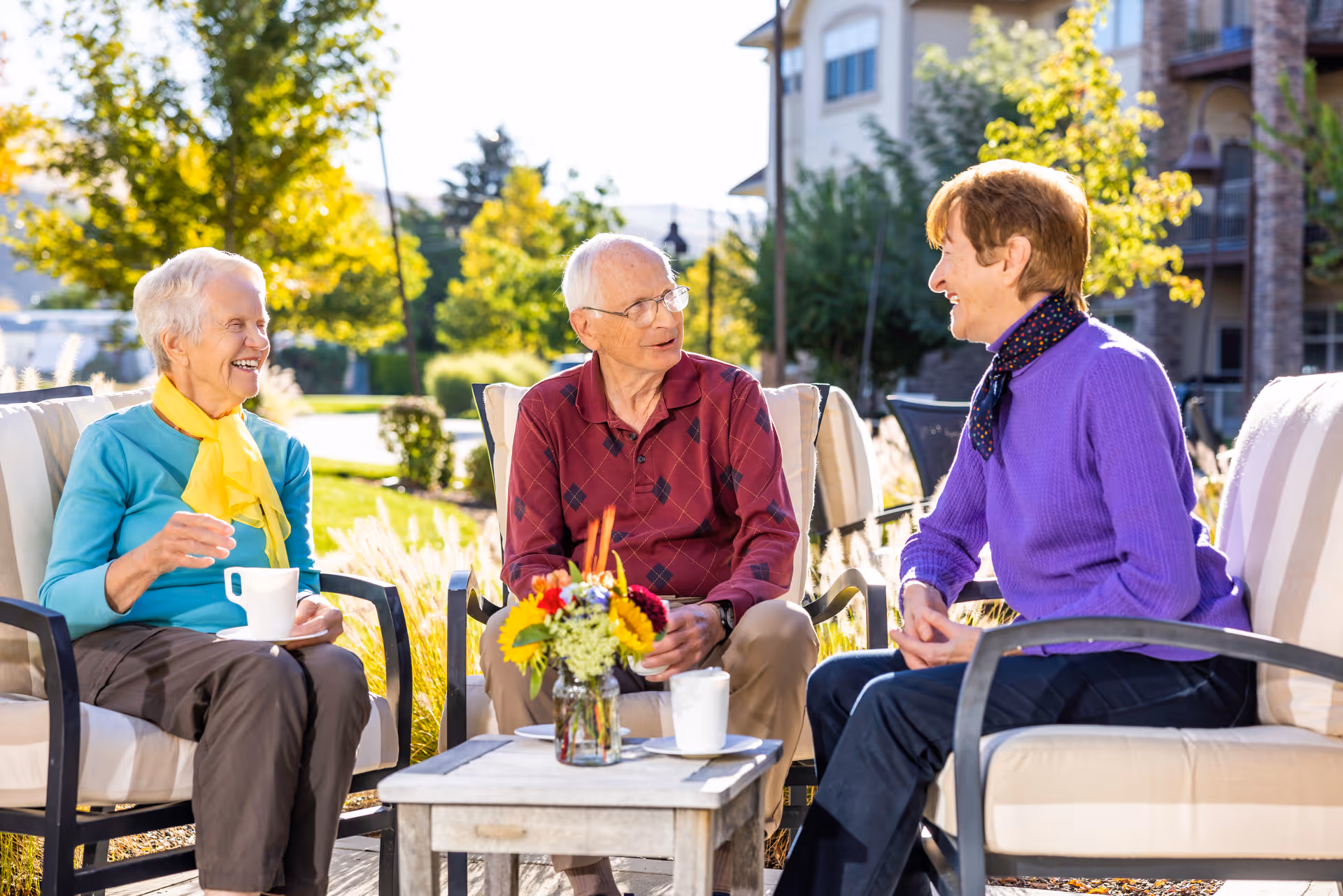 Three elderly people sitting outdoors on cushioned patio chairs around a small table with a flower vase, enjoying a sunny day and having a conversation with coffee cups in hand. Trees and a building are visible in the background.