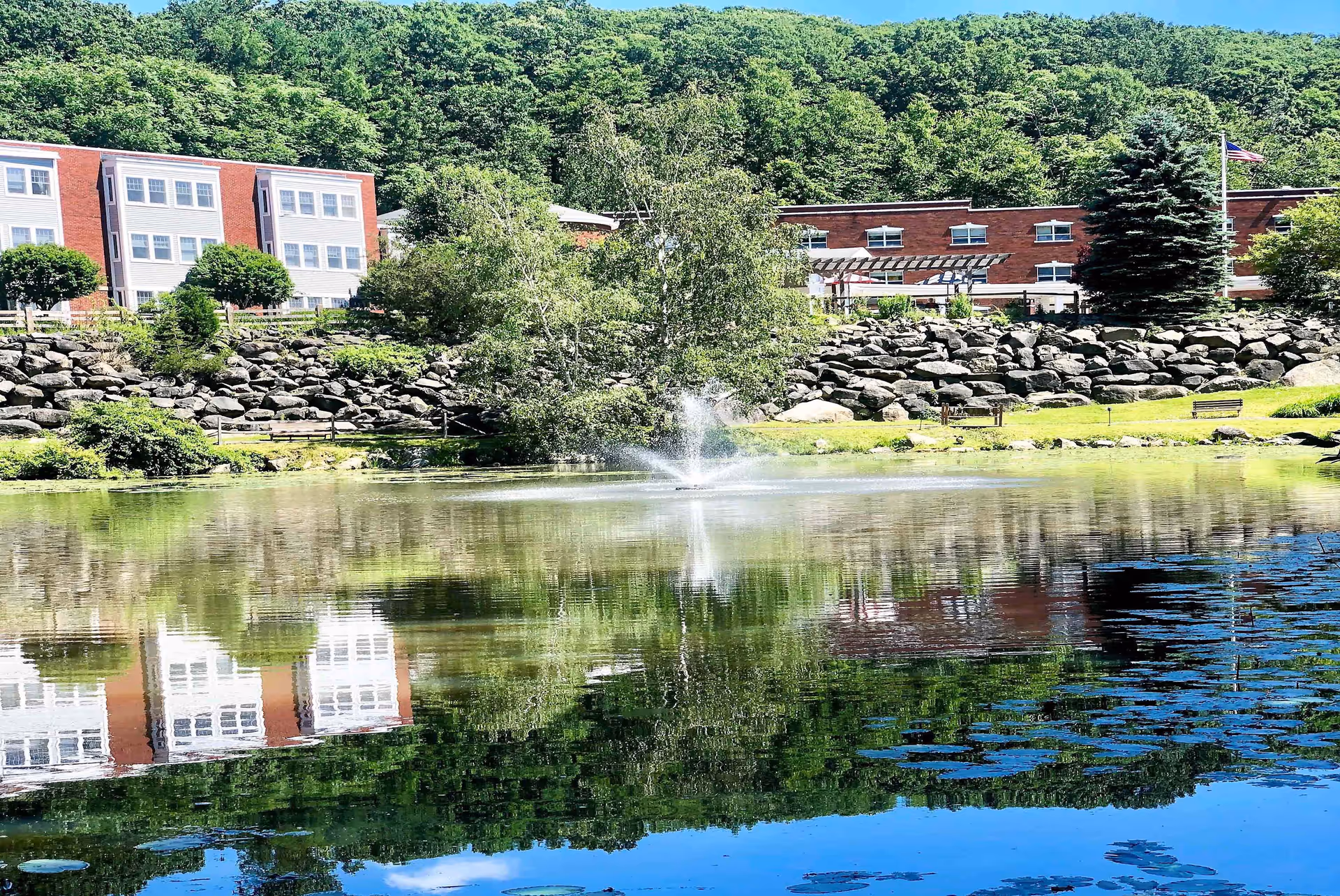 A pond with a central fountain reflecting a red-brick senior living building and tree-covered hillside behind it.