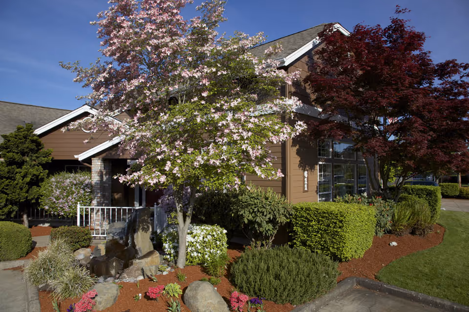 Well-landscaped front exterior of a brown residential building with flowering trees, shrubs, and a small rock garden.