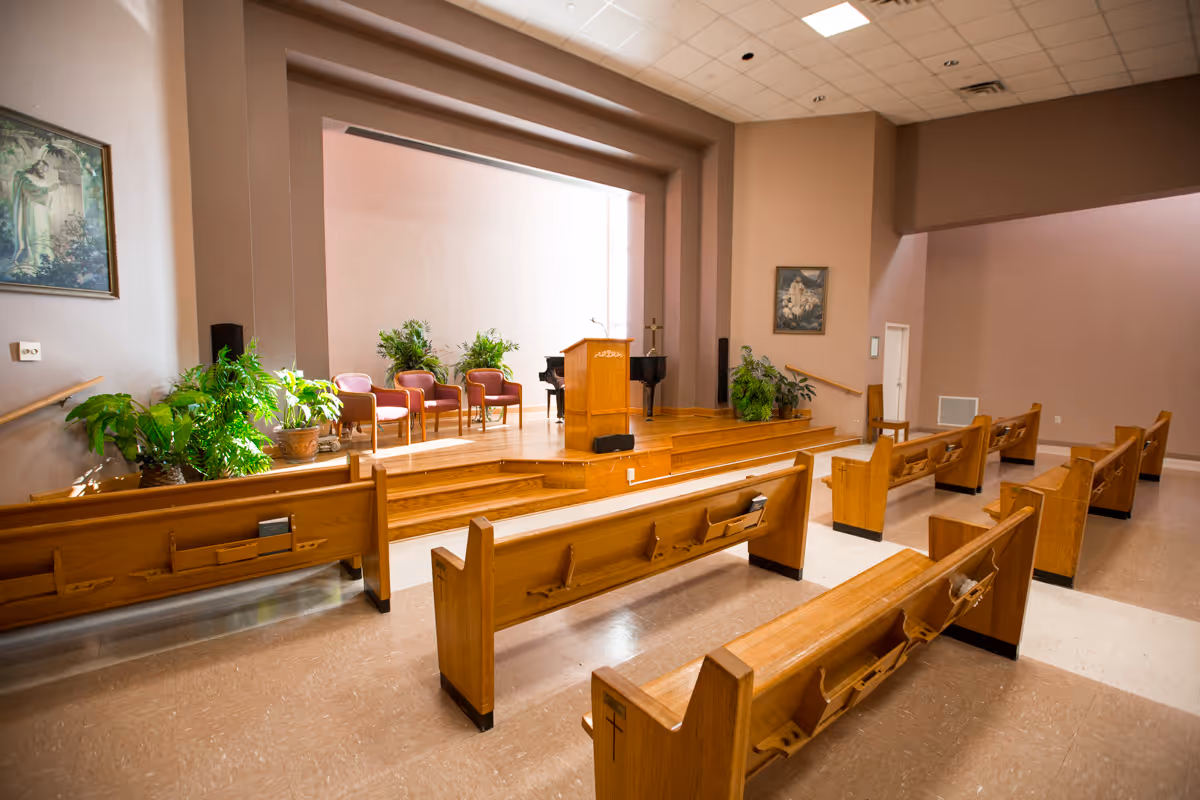 Interior view of a chapel or small auditorium with wooden pews arranged facing a raised stage. The stage has a wooden podium, several chairs, a piano, and potted plants. The walls are painted light brown, and there are framed pictures hanging on the walls.