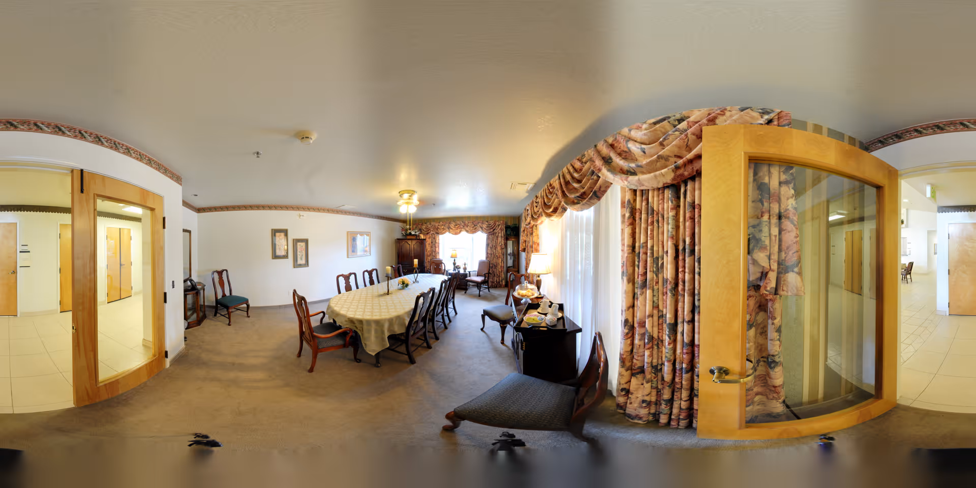 A spacious dining room with a long table covered with a white tablecloth and surrounded by wooden chairs. The room has beige carpet, floral curtains on large windows, and a sideboard with a lamp and tea set. There are framed pictures on the walls and a ceiling light fixture. The room is connected to a hallway with tiled flooring through wooden doors with glass panels.