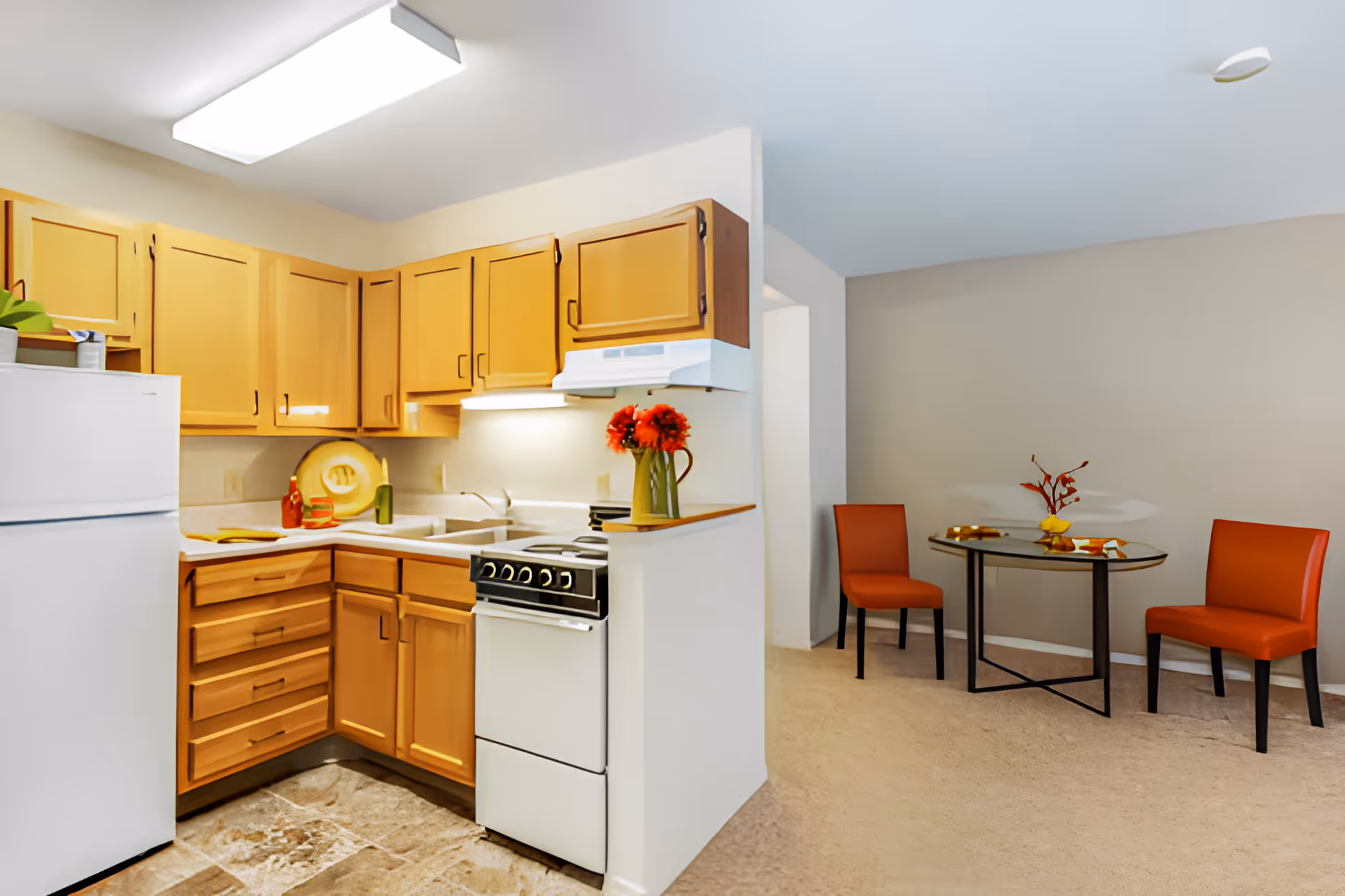 Small open kitchen with wooden cabinets, white appliances, and a nearby dining nook with a round table and two orange chairs.