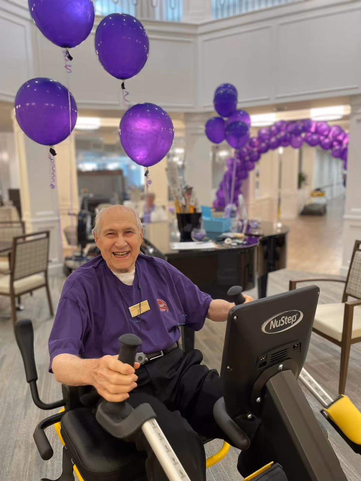 An elderly man wearing a purple shirt is smiling while using a NuStep exercise machine in a well-lit indoor space decorated with purple balloons and a purple balloon arch in the background.
