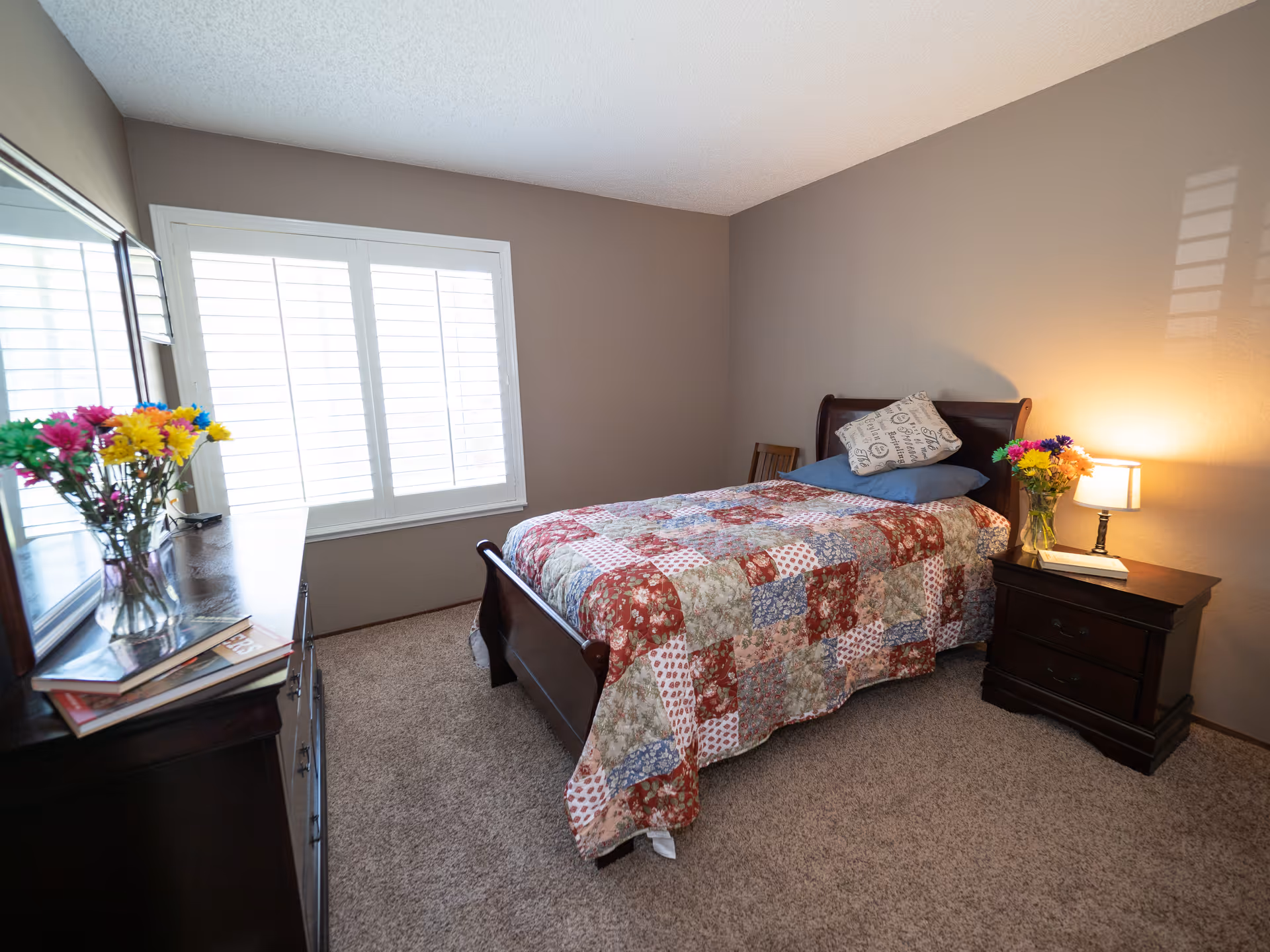 A neatly furnished bedroom with a single bed in a patchwork quilt, nightstand with a lamp and flowers, and a dresser by a shuttered window.