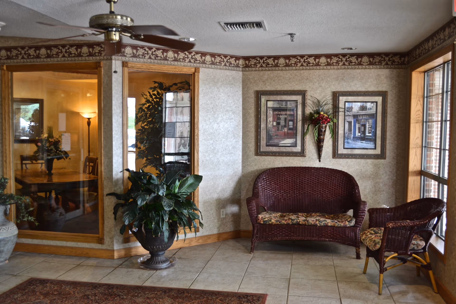 A cozy sitting area with a wicker loveseat and chair, both with floral cushions, placed near a large window. The walls are decorated with two framed pictures and a wall-mounted vase with flowers. There is a large potted plant on the tiled floor and a ceiling fan above. Through glass windows, a dining table with chairs and a floor lamp can be seen in an adjacent room.