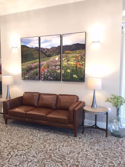 A cozy seating area with a brown leather couch, two side tables each holding a lamp with a white shade, and a large glass vase with green foliage on the floor. Above the couch is a three-panel wall art depicting a colorful mountain landscape with wildflowers. The floor is covered with a patterned carpet.