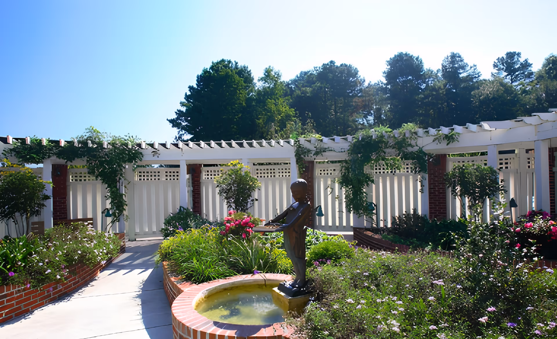 A sunny outdoor garden area at Cypress Glen Retirement Community featuring a circular brick planter with a small water fountain and a bronze statue of a child playing a violin. The garden is lush with various green plants and colorful flowers, surrounded by white wooden trellises with climbing vines and tall trees in the background.