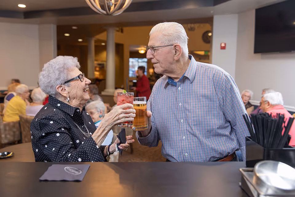 Two elderly residents smiling and clinking drinks across a bar in a communal dining/lounge area.