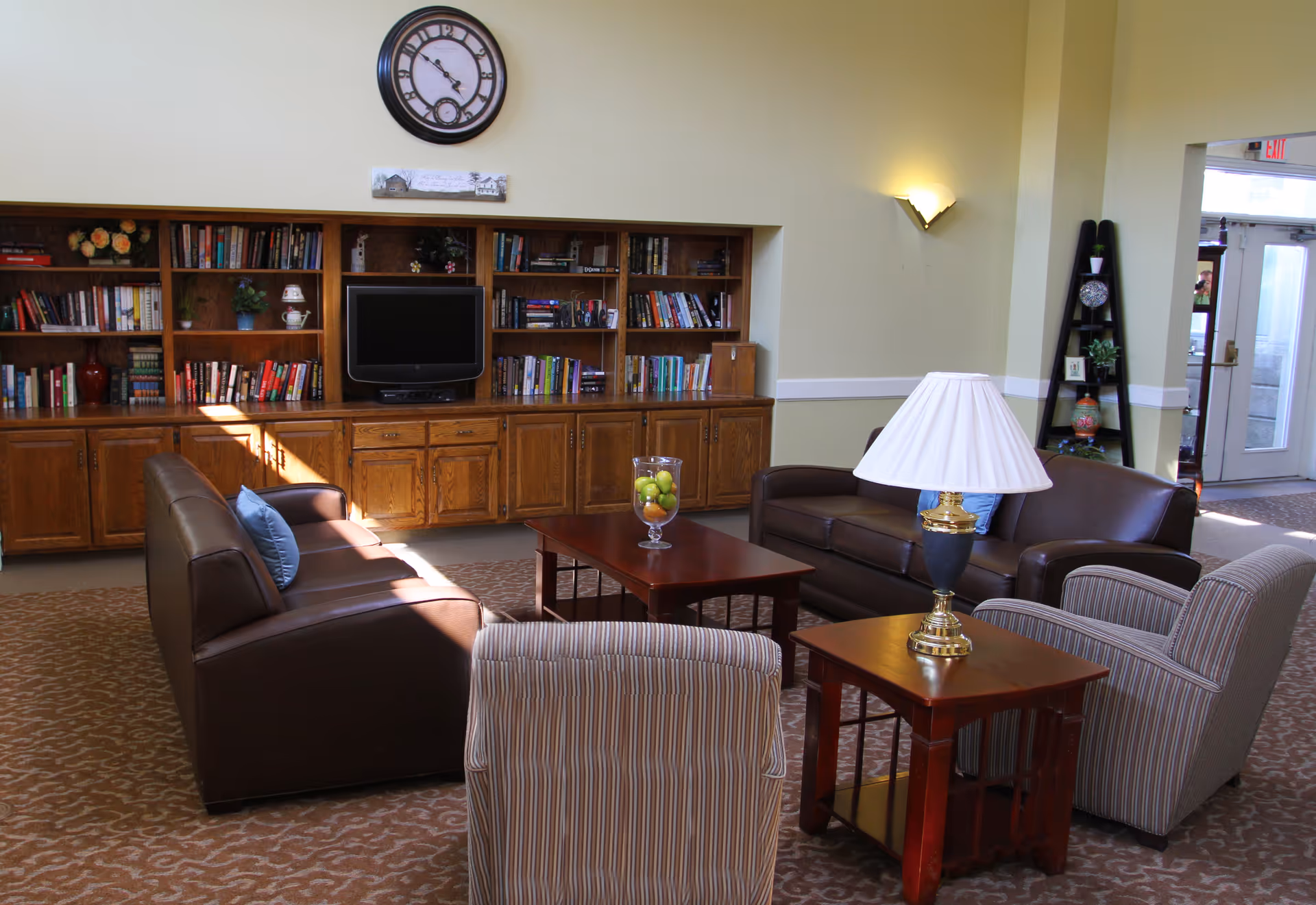 Comfortable senior living lounge with sofas and armchairs arranged around a coffee table, built-in bookshelves with a TV, and a large wall clock.