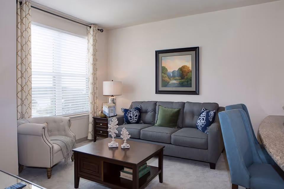 A cozy living room with a gray sofa adorned with green and blue patterned pillows, a beige armchair with a gray throw blanket, a wooden coffee table with decorative items, a side table with a lamp, and a large window with white blinds and patterned curtains. A framed landscape painting hangs on the wall above the sofa.