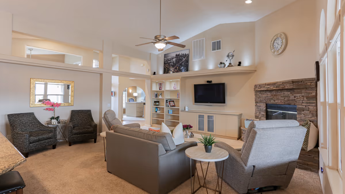 A cozy living room in a senior care home featuring a gray sofa, two gray armchairs, a stone fireplace, a wall-mounted TV, built-in shelves with decorative items, a ceiling fan, and a large wall clock. The room has beige walls and carpeted floors, with a mirror and a small table with flowers near the seating area.