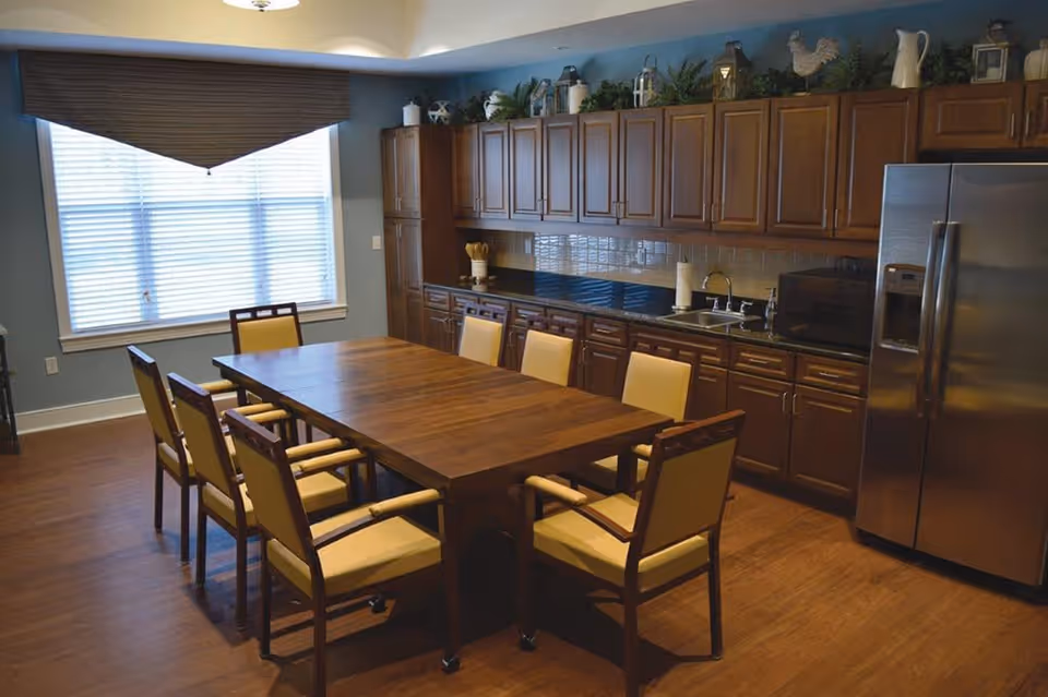 A kitchen dining area with a large wooden table surrounded by eight cushioned chairs. The room features wooden cabinets along one wall, a stainless steel refrigerator, a sink, and various decorative items on top of the cabinets. A large window with blinds allows natural light into the space.