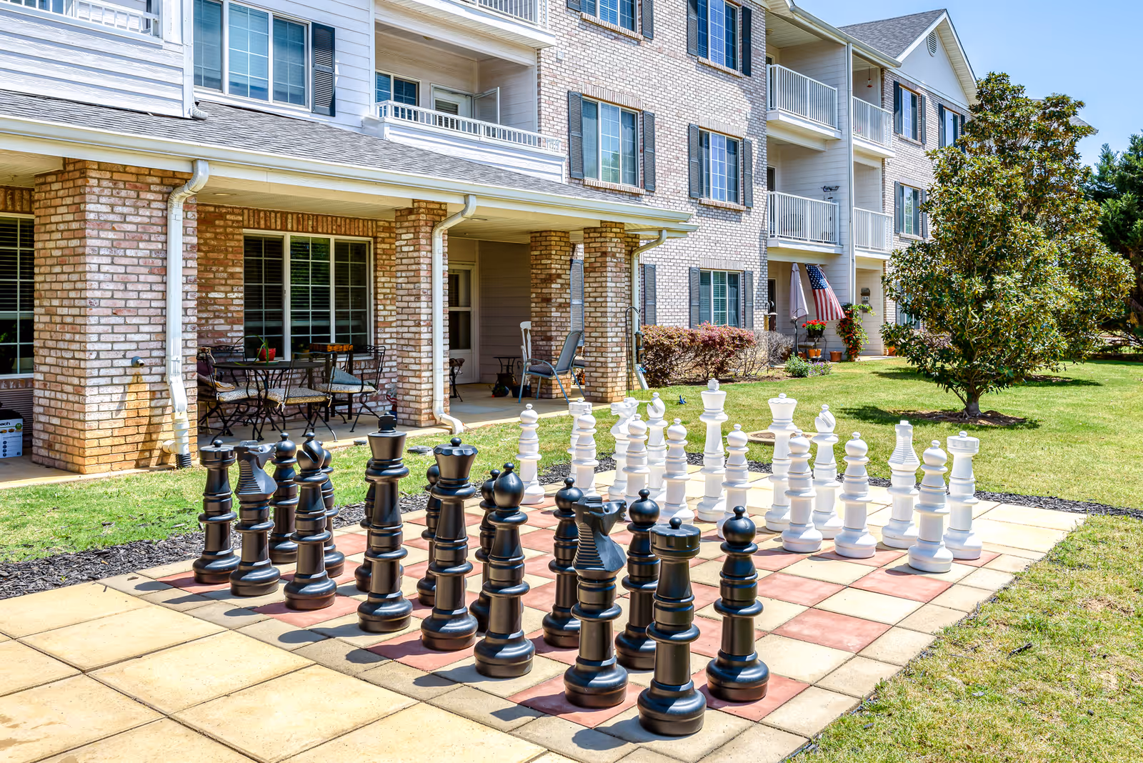 Large outdoor chessboard with oversized black and white pieces on a tiled patio in front of a multi-story brick senior living building.