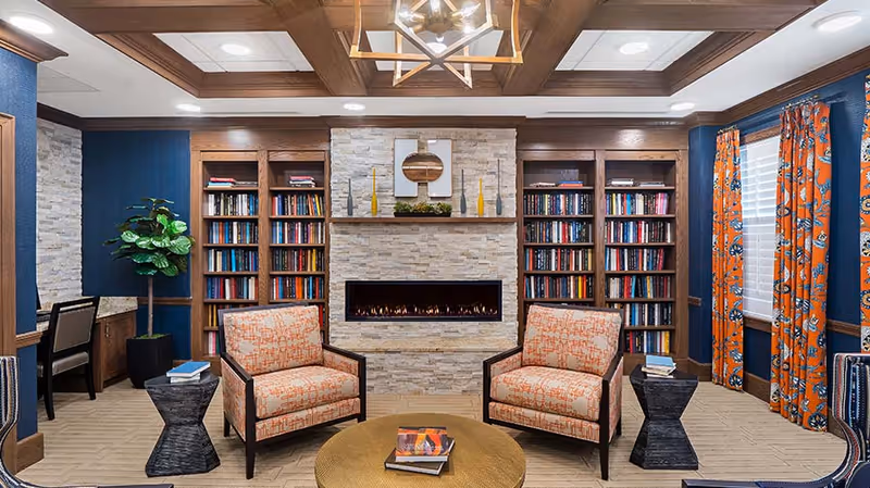 A cozy living room area featuring two patterned armchairs with orange upholstery facing a modern electric fireplace set in a stone wall. The fireplace is flanked by built-in wooden bookshelves filled with books. A round coffee table with books on it is in front of the chairs. The room has blue walls, orange patterned curtains on the window, a wooden coffered ceiling with a geometric chandelier, and a potted plant in the corner.