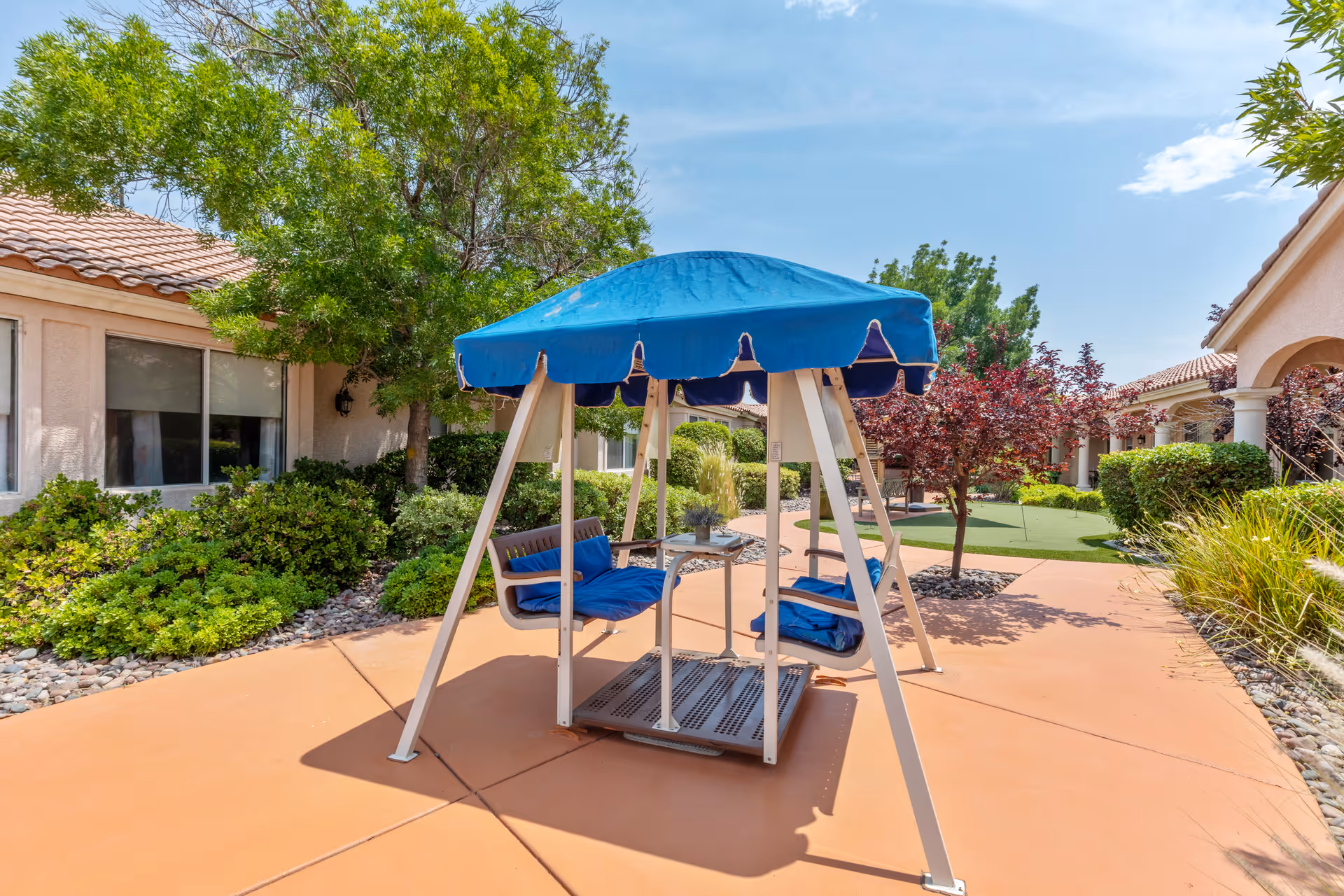 Outdoor patio area at a senior living facility with a blue canopy swing seat, surrounded by greenery, trees, and shrubs. The area has a paved walkway and is adjacent to buildings with tiled roofs and large windows.