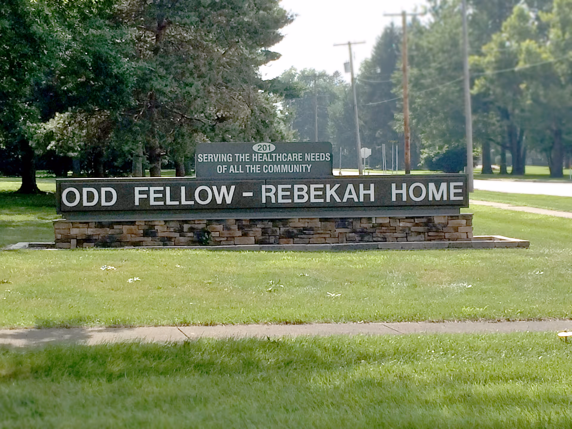 Stone entrance sign reading 'Odd Fellow - Rebekah Home' on a grassy lawn with trees and a road behind it.