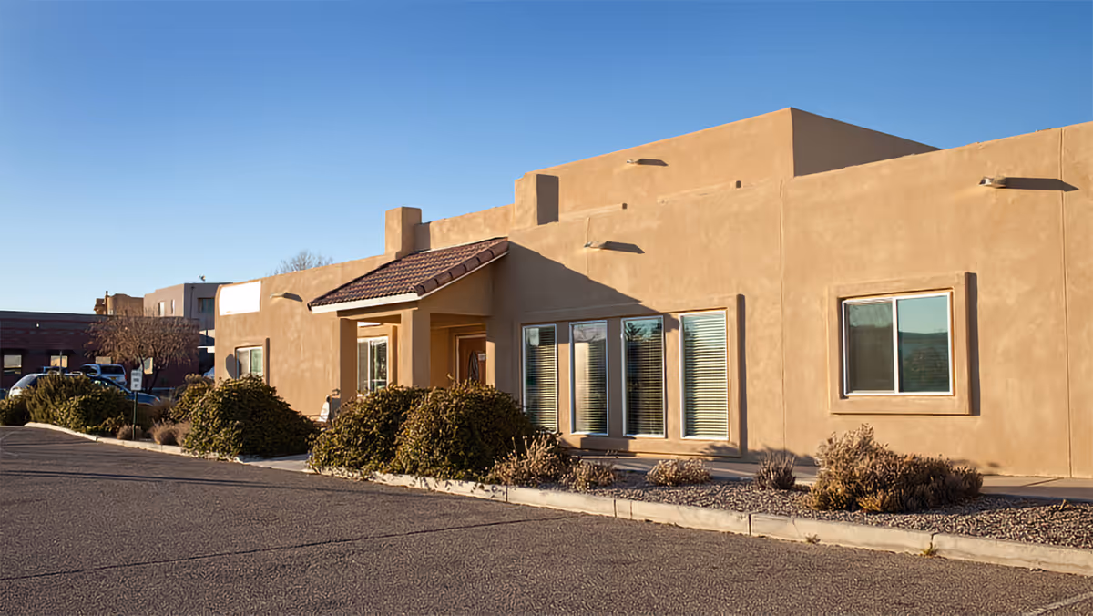 Exterior view of a single-story building with adobe-style architecture, beige stucco walls, and a tiled roof over the entrance. There are several windows with blinds and landscaping with bushes and gravel along the front. The sky is clear and blue.