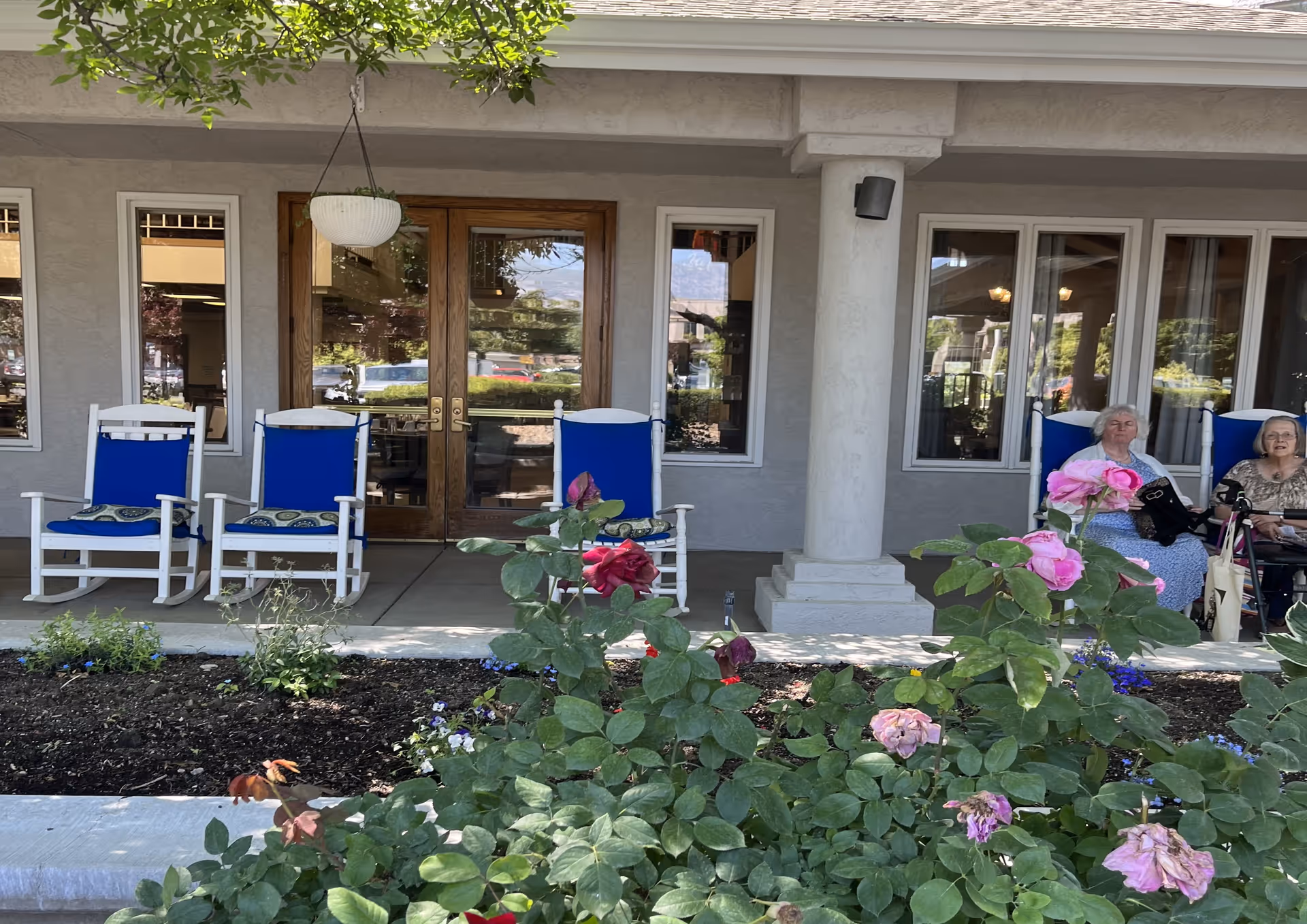 Outdoor patio area at Solista Orem with four white rocking chairs that have blue cushions. Two elderly women are seated on the right side, one in a wheelchair. There are rose bushes with pink and red flowers in the foreground, and a hanging white planter above the chairs. The building has beige walls, wooden double doors, and multiple windows.