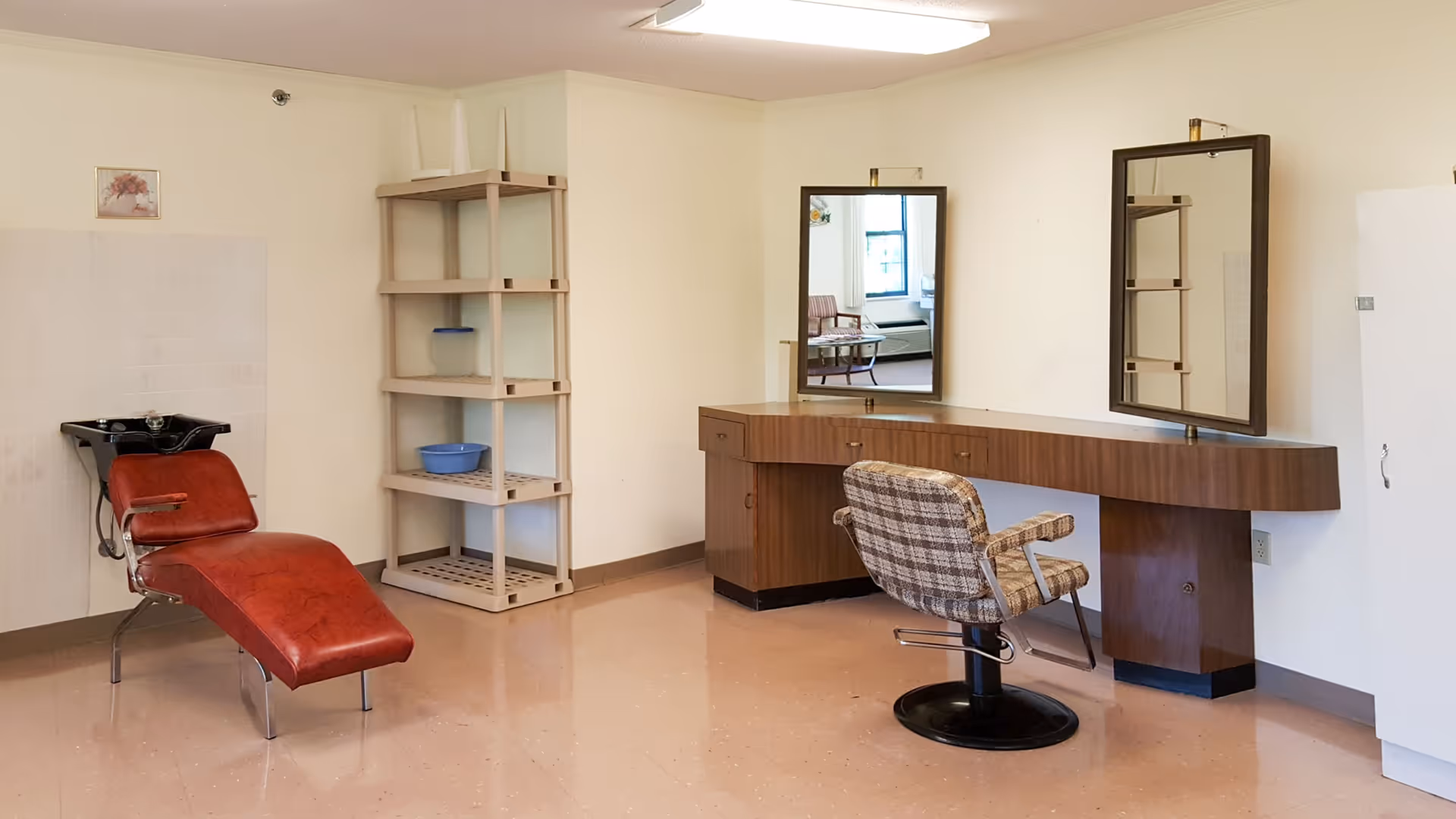 Interior salon area with a red reclining shampoo chair, a plaid styling chair in front of a long vanity with mirrors, and shelving against the wall.