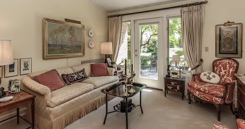 A cozy living room with a beige sofa adorned with red and patterned cushions, a glass coffee table with decorative items, a red upholstered armchair with a white pillow, side tables with lamps, framed artwork on the walls, and glass doors leading to an outdoor patio area with greenery.