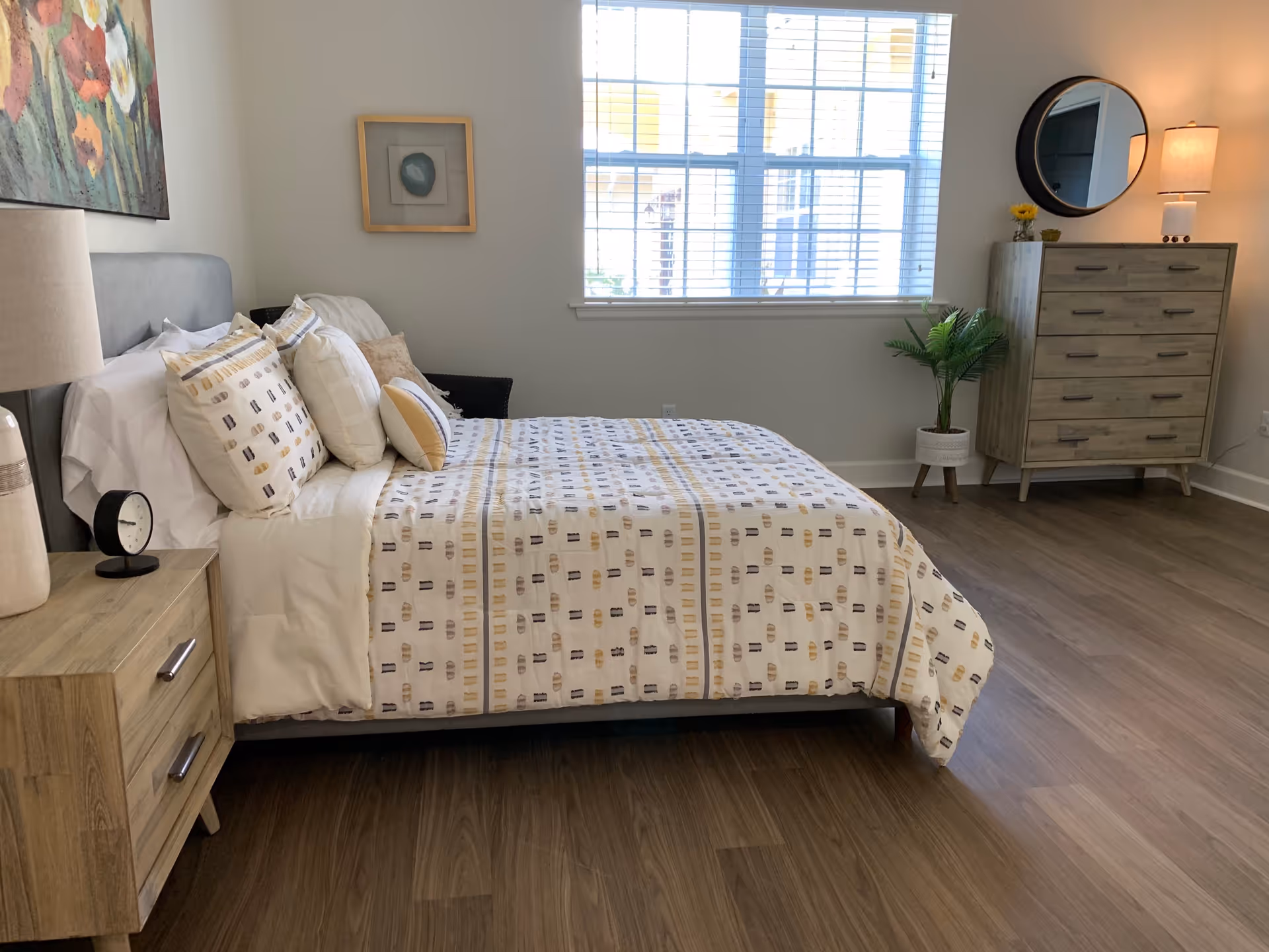 A neatly made bed with patterned bedding and multiple pillows in a bright bedroom. The room features wooden flooring, a wooden nightstand with a lamp and clock, a dresser with a round mirror and lamp, a potted plant, and a large window with blinds letting in natural light.