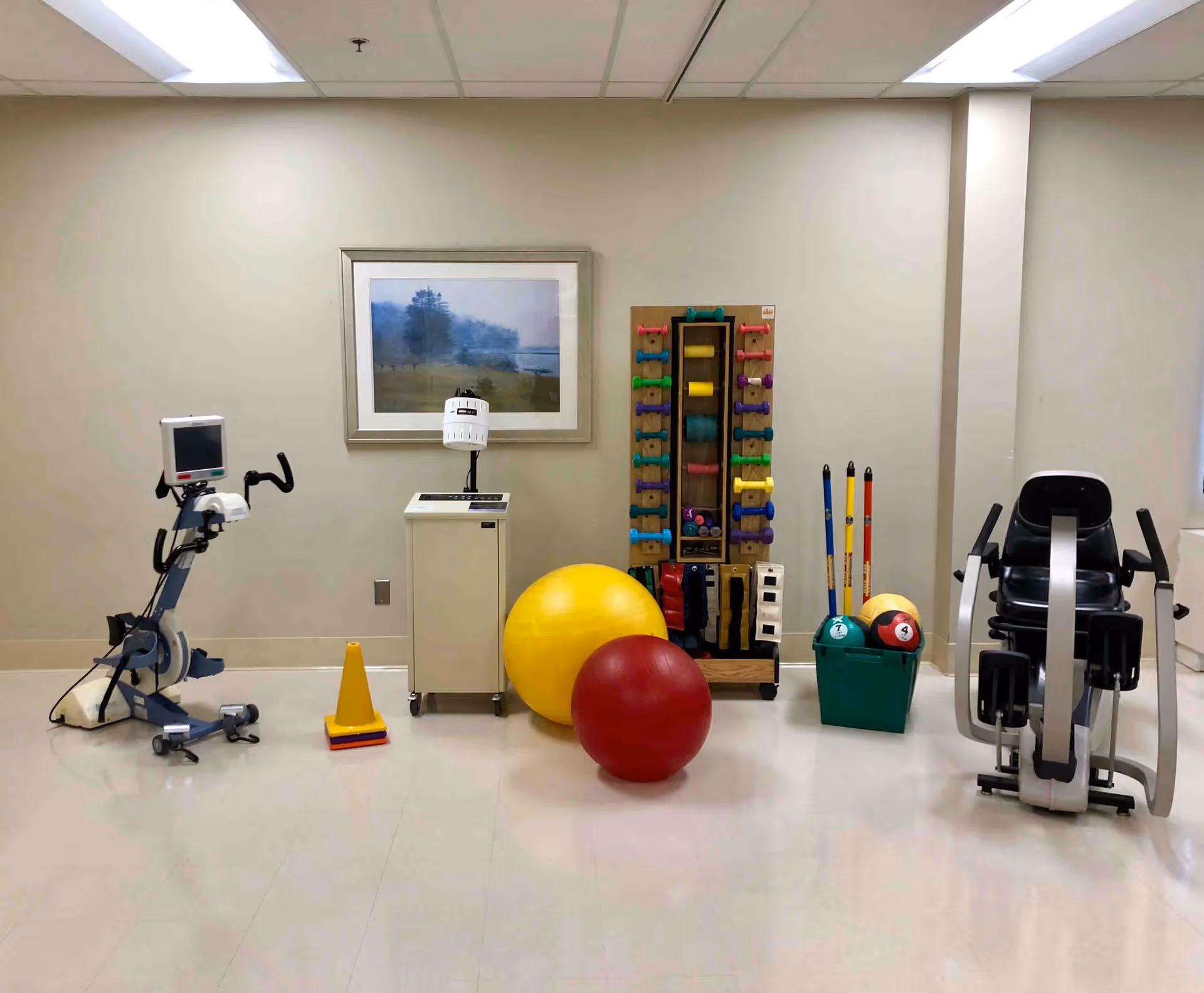 A rehabilitation exercise room with various fitness equipment including a stationary bike, exercise balls, colorful dumbbells on a rack, weighted balls, and other physical therapy tools. The room has beige walls, a framed landscape picture, and a tiled floor.