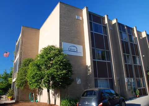 Exterior view of a multi-story senior living facility building with a sign that reads 'Epoch Senior Living'. There is a tree and some greenery in front of the building, a parked car, and an American flag on the left side. The sky is clear and blue.